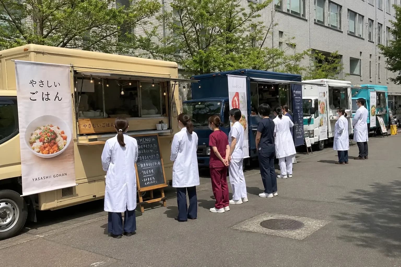 病院の敷地内に複数のキッチンカーが並び、白衣のスタッフが並んでいる写真